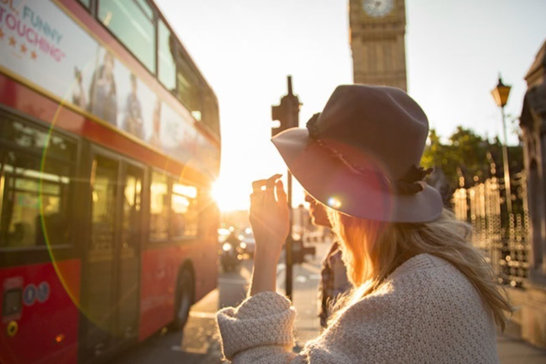 A woman wearing a hat stands in the sunset in front of a red double-decker bus and Big Ben in London.