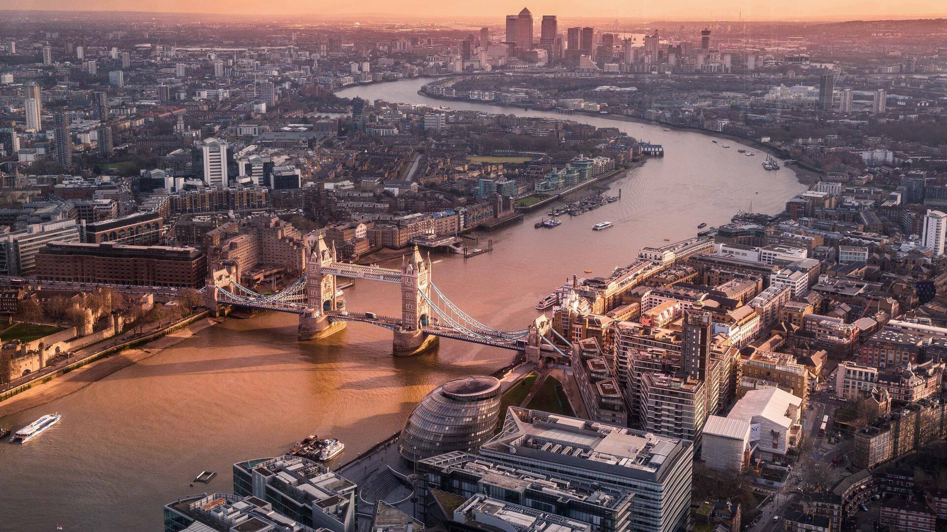 This is a panoramic aerial view of London, featuring the River Thames winding through the city, with the iconic Tower Bridge prominently visible, and the city skyline in the background.