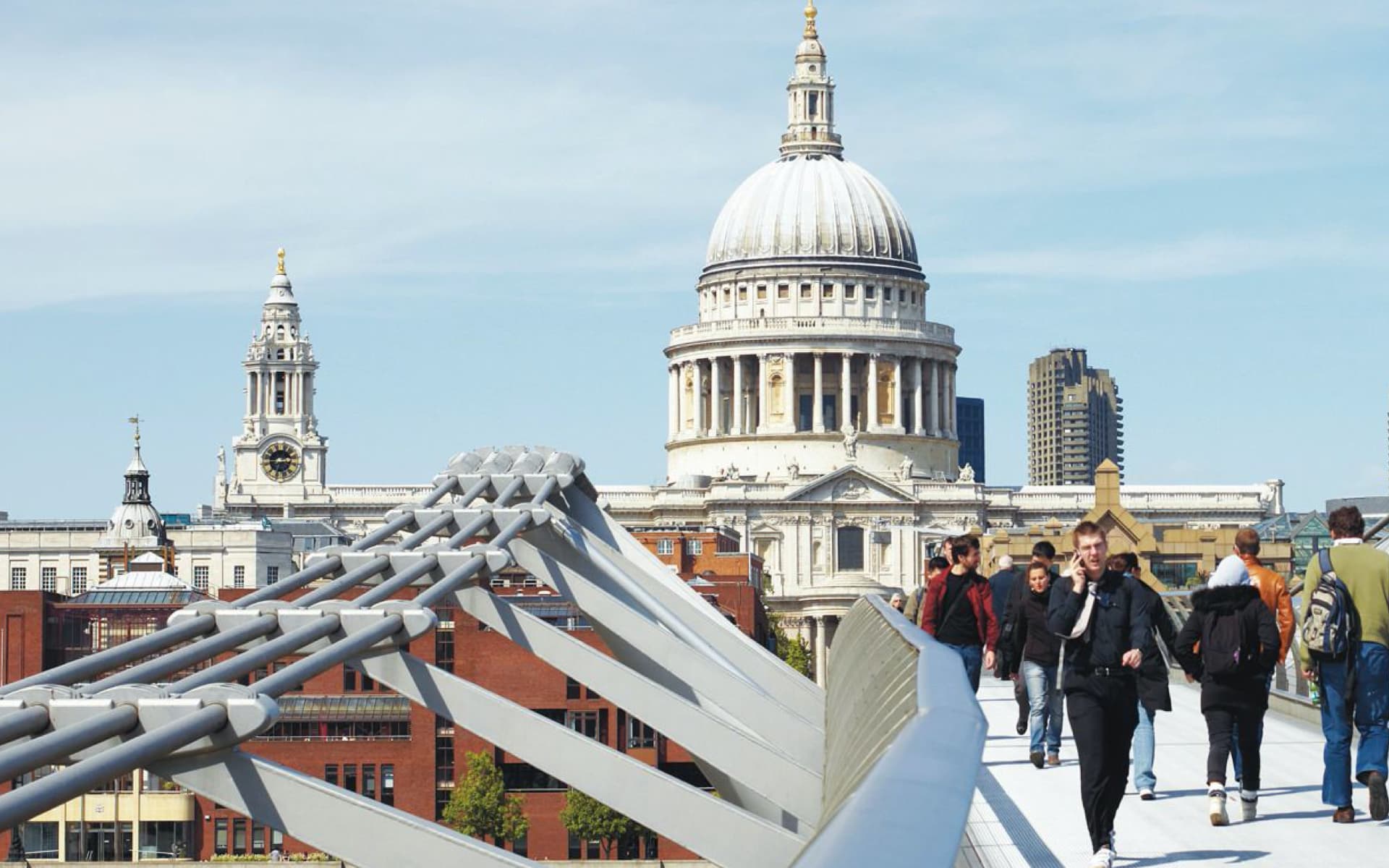 This image shows the Millennium Bridge in London, with St. Paul's Cathedral visible in the background. People are walking across the bridge on a sunny day.