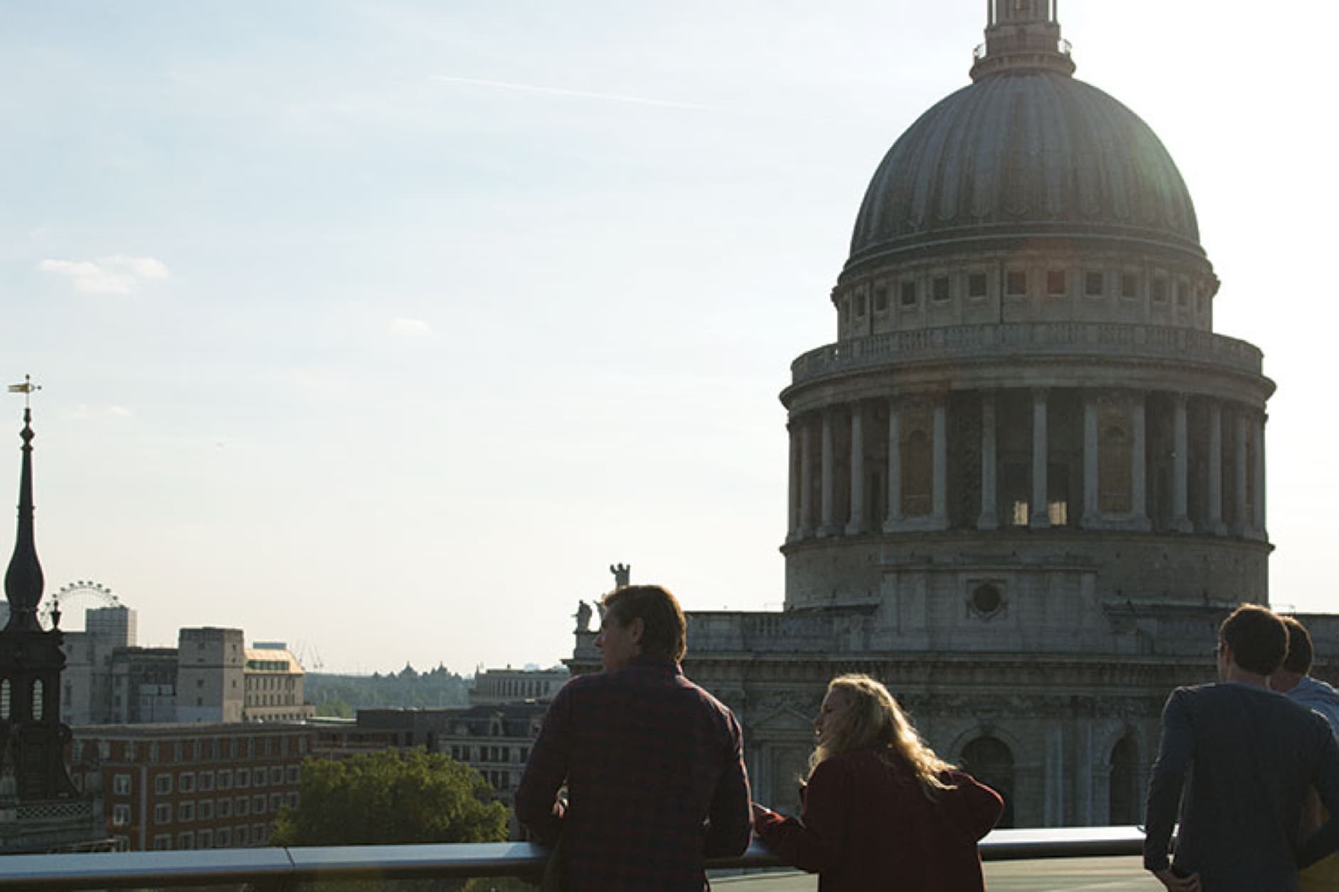 Several people stand on a rooftop, looking out at St. Paul's Cathedral in London. The sun is shining on the iconic building.