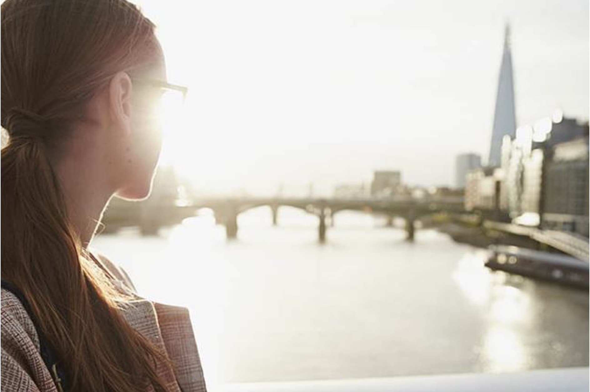 A woman with her hair in a ponytail looks out over a sunny river with a bridge and the London skyline, including the distinctive Shard building, in the background.