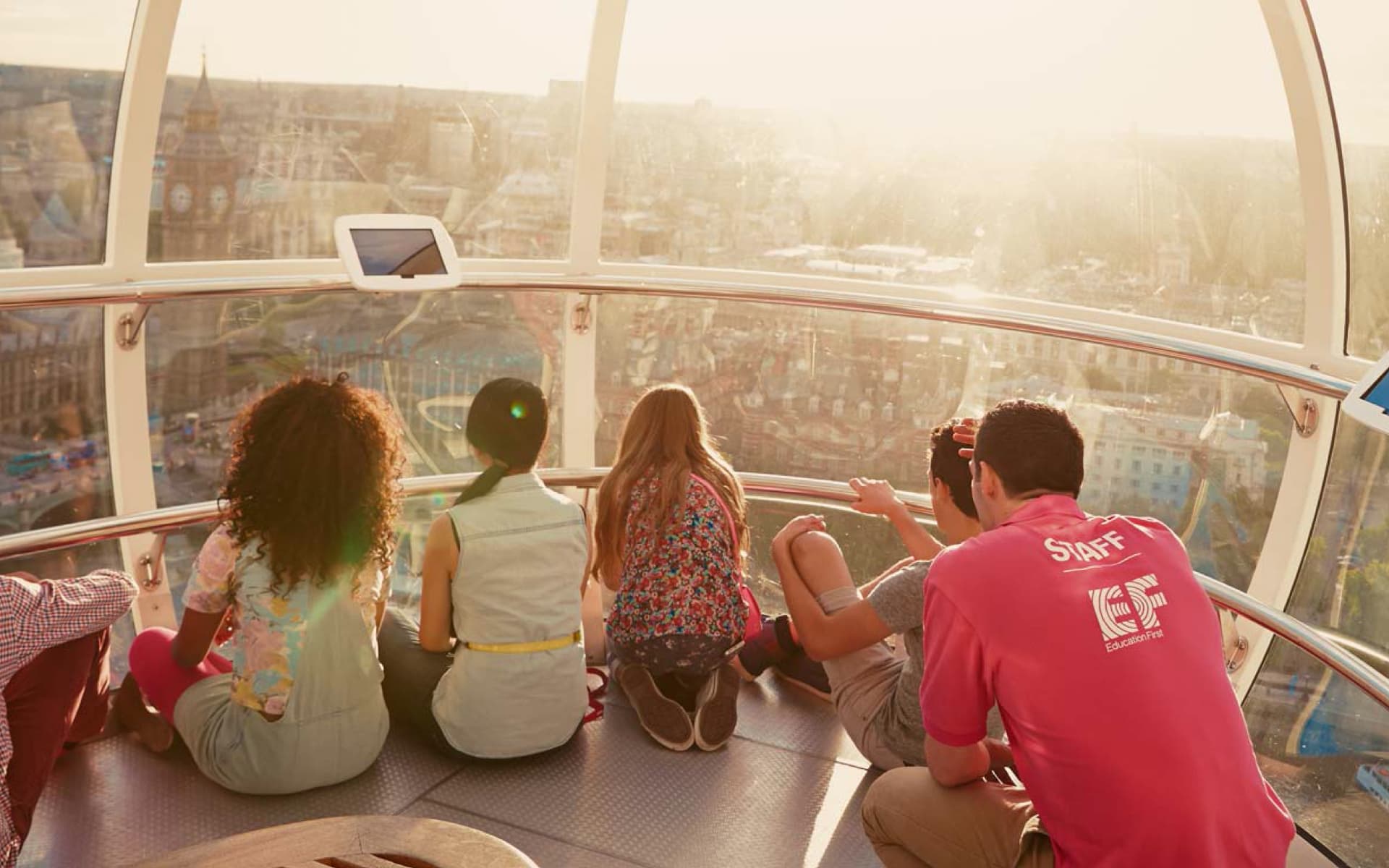 A group of five young people are sitting inside a capsule, looking out at a city during sunset.