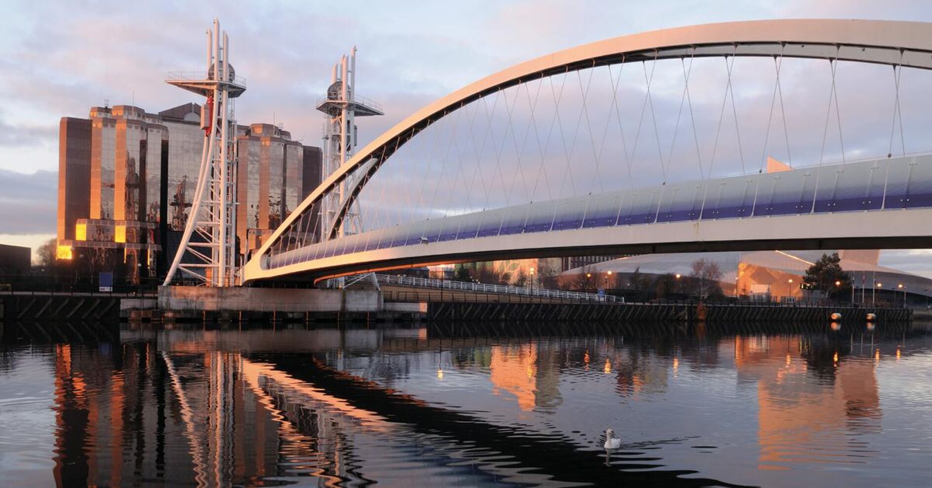 A modern arched bridge spans a calm river, with buildings and towers in the background reflecting in the water. The scene is illuminated by the rising sun.