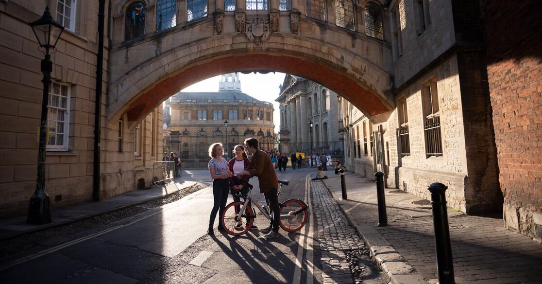 A group of three people with a bicycle stand on a cobblestone street under an arched stone bridge. The architecture suggests a historic European city, possibly Oxford, with warm sunlight casting long shadows.