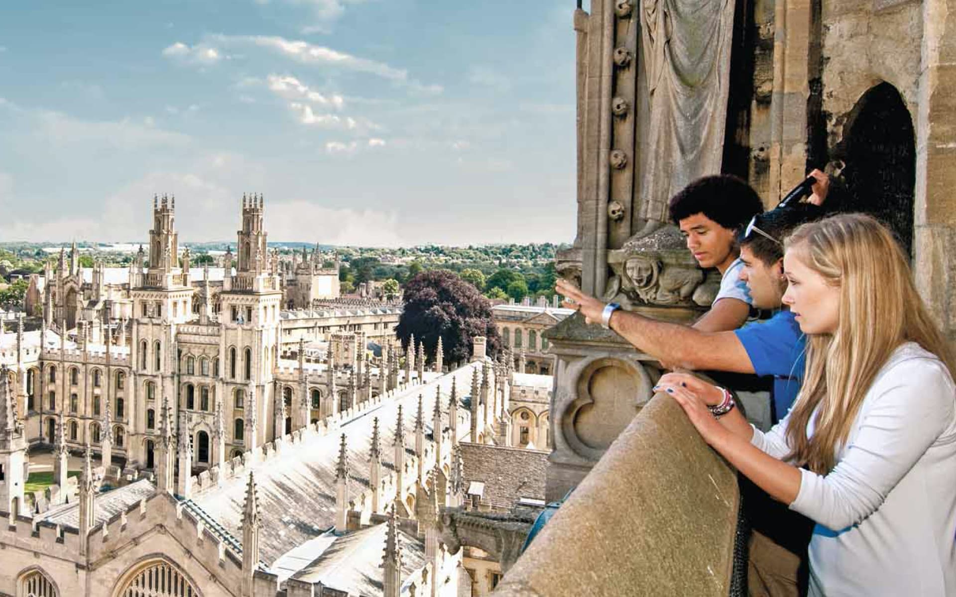 Three young people are enjoying the view from a high balcony, overlooking a historic city with numerous spires and buildings.
