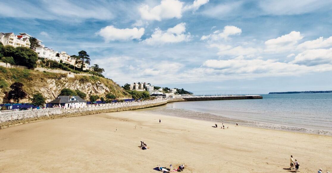A scenic view of a sandy beach with a few people enjoying the sun and sea. Buildings are visible on a hill overlooking the coastline, with a pier extending into the calm blue water under a partly cloudy sky.