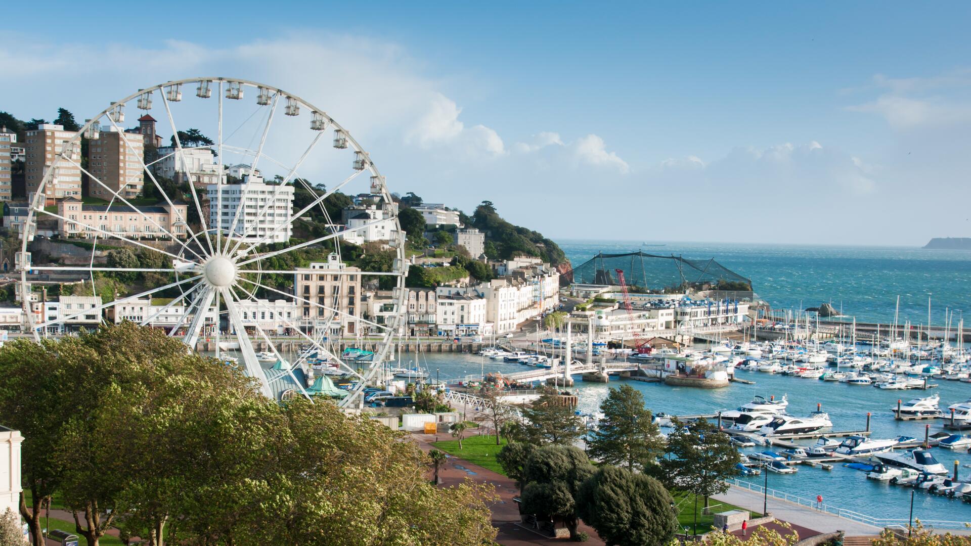 This image shows a scenic view of a coastal town with a large Ferris wheel overlooking a bustling harbor filled with numerous boats and yachts under a clear blue sky.