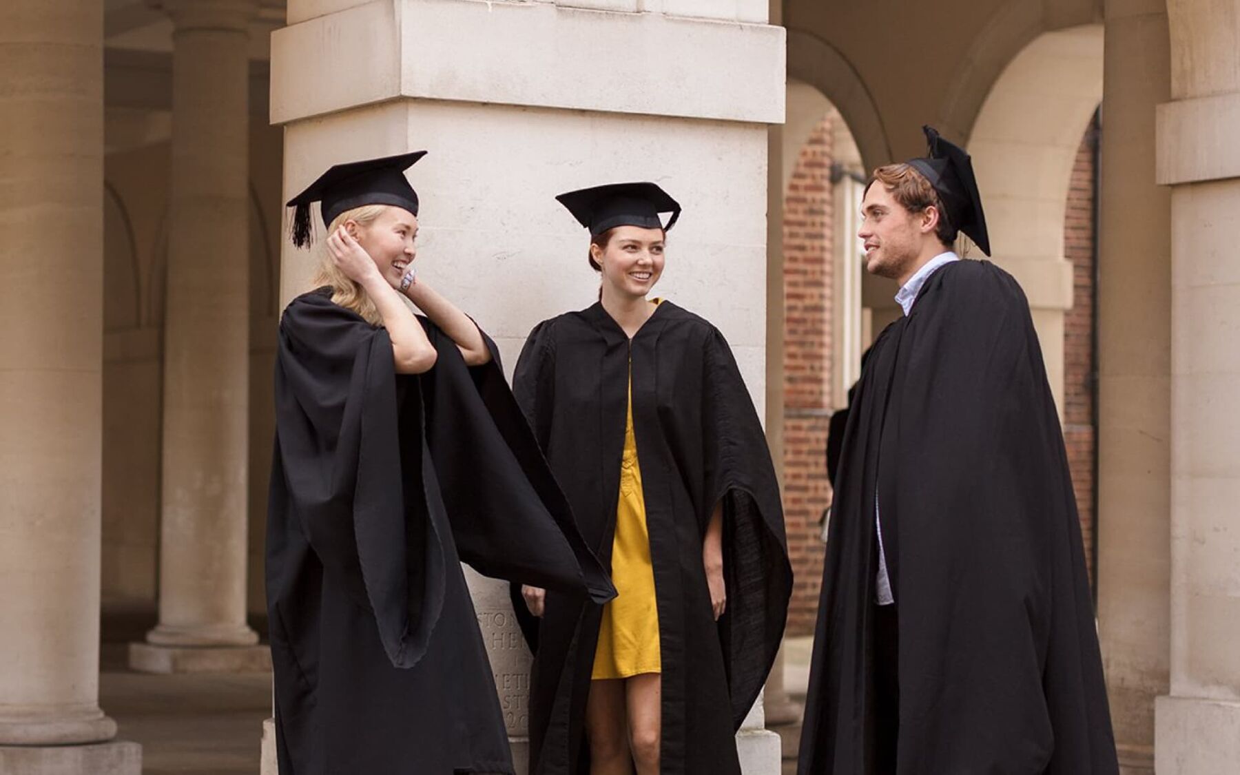 Three students in graduation gowns and caps are chatting and smiling in front of a colonnaded building, signifying a successful academic achievement.