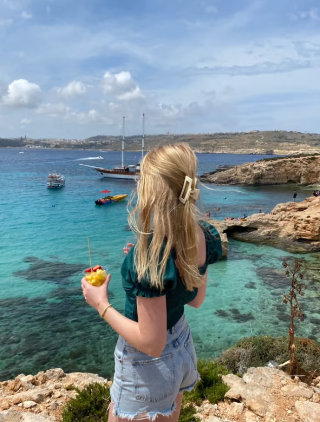 A young woman with blonde hair, wearing denim shorts and a green top, enjoys a refreshing drink while admiring a breathtaking turquoise sea with boats and rocky shores under a clear blue sky.