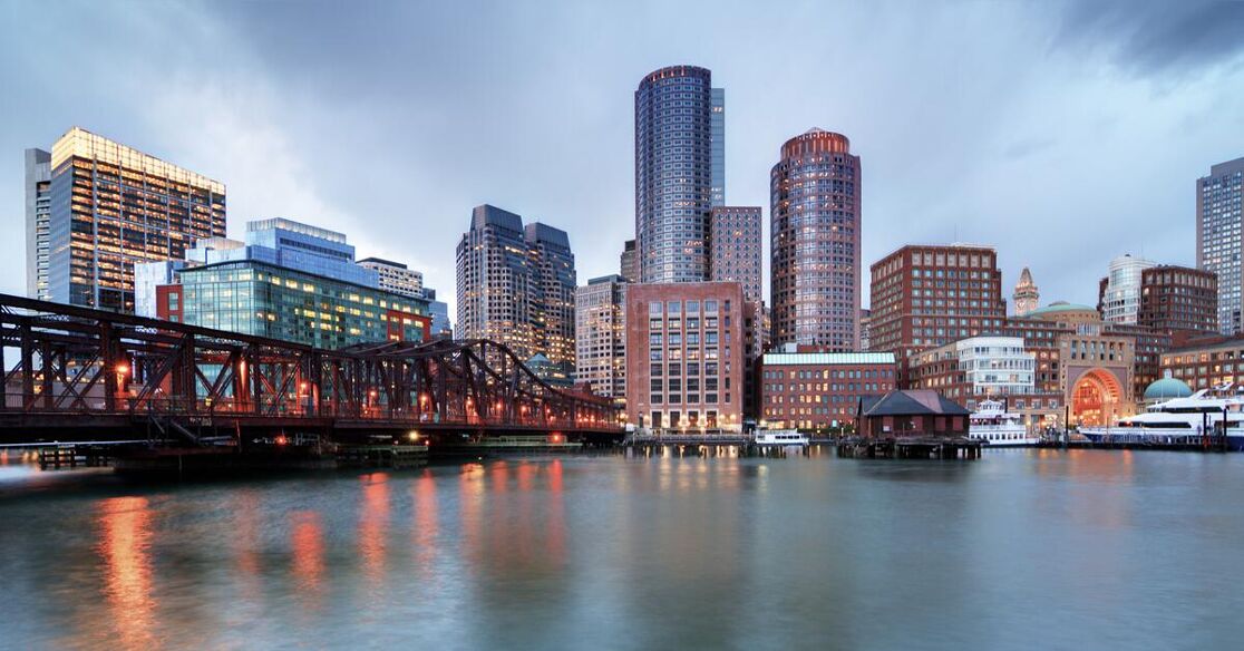 The Boston skyline is visible across the Charles River, with a bridge in the foreground, illuminated during twilight.