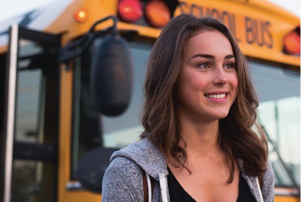 A young woman with brown hair, wearing a grey hoodie, smiles in front of a school bus. She has a thoughtful expression.