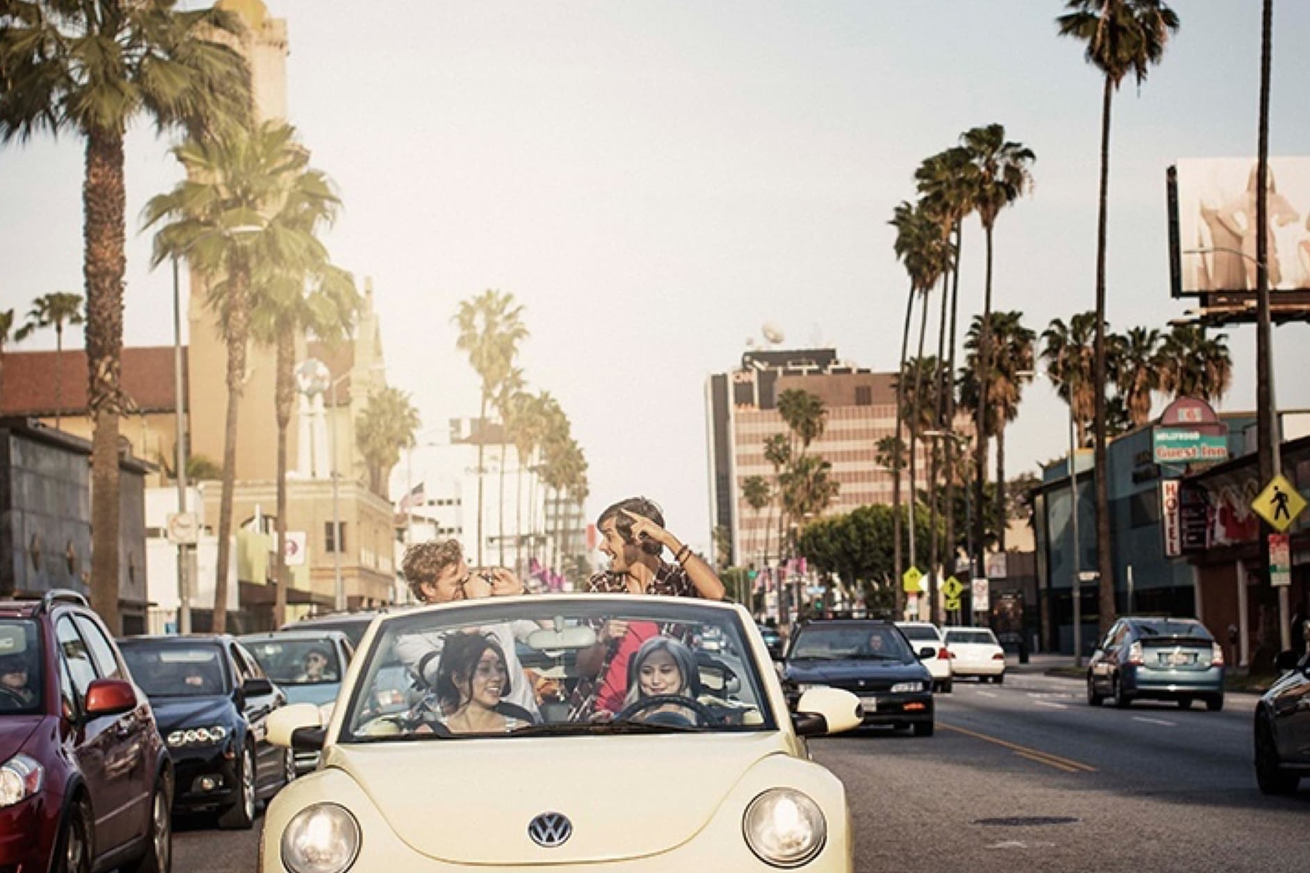 A group of friends are cruising in a convertible down a sunny city street, surrounded by palm trees and other cars. They appear to be having a great time.