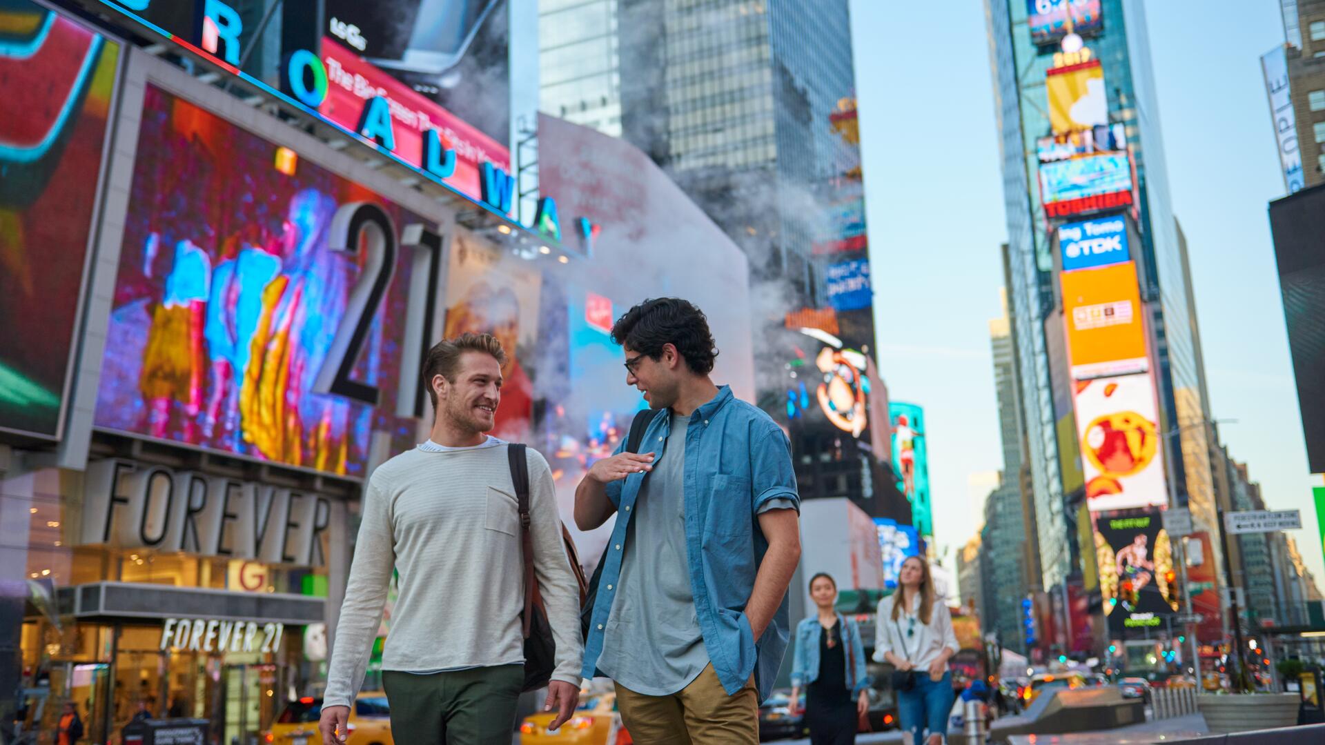 Two men walk through a bustling street in New York City, passing by large billboards and skyscrapers that make up the city's famous skyline.
