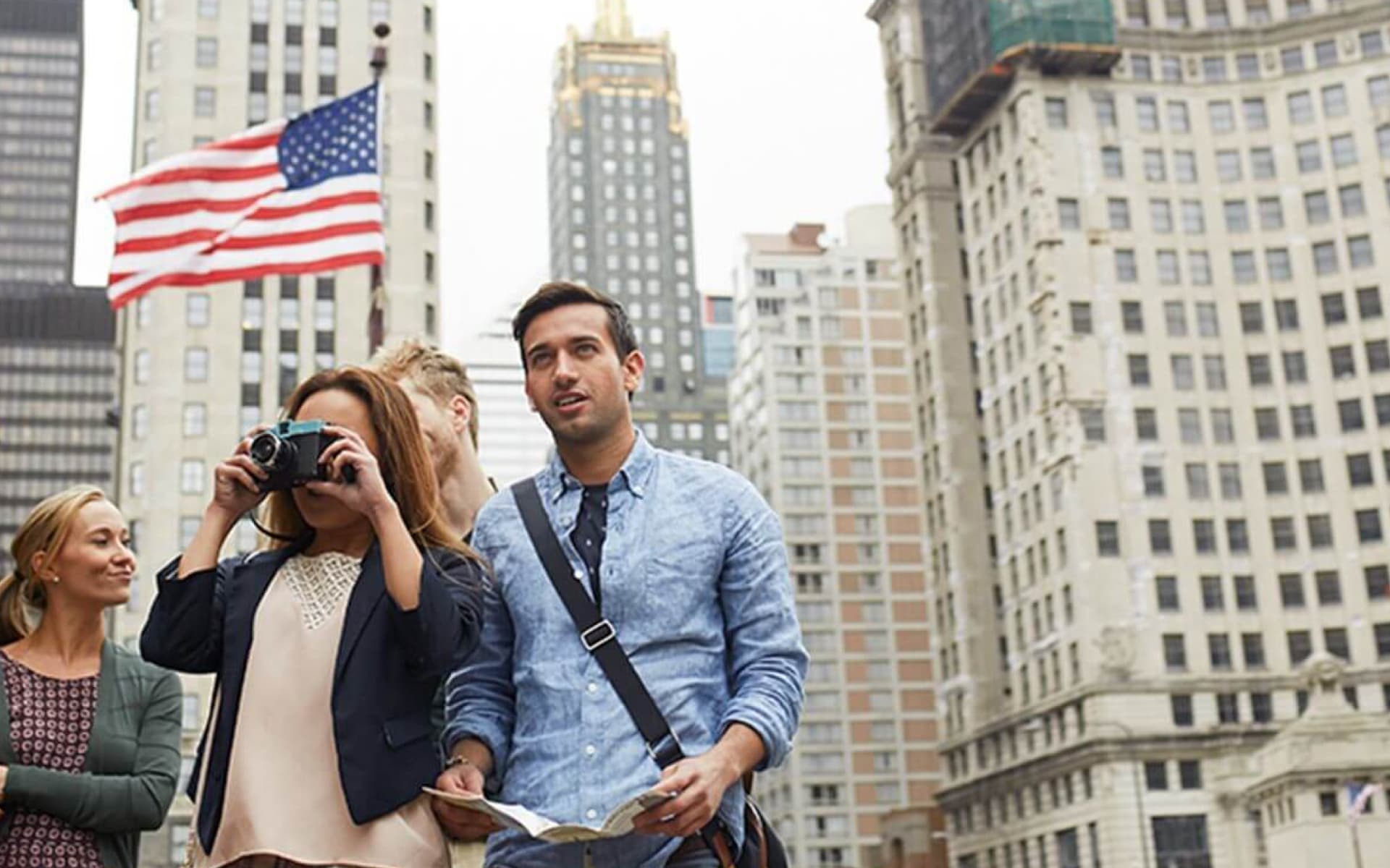 A group of tourists are exploring a city, with one woman taking photos with a vintage camera. An American flag waves in the background, suggesting a location in the United States.