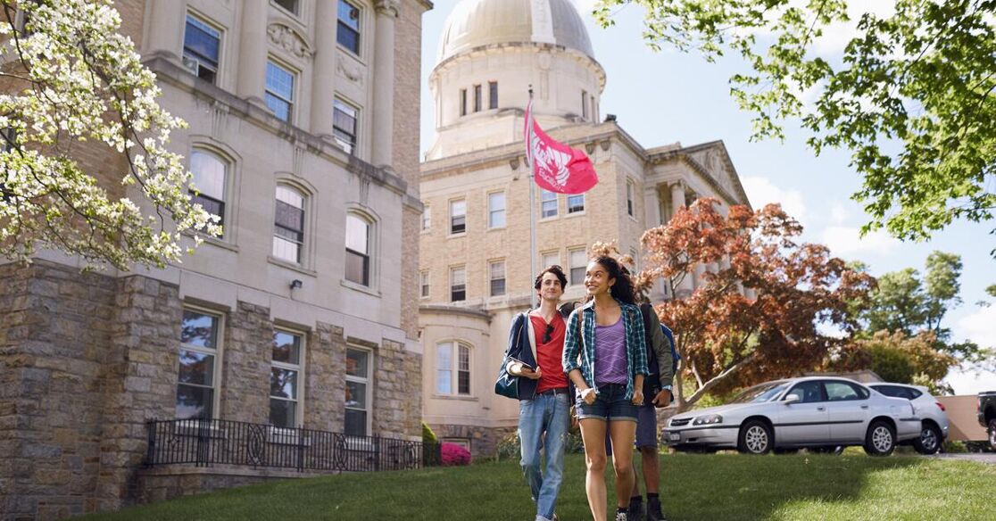 Two students walk across a sunny campus, passing by a historic building featuring a dome and a flag.