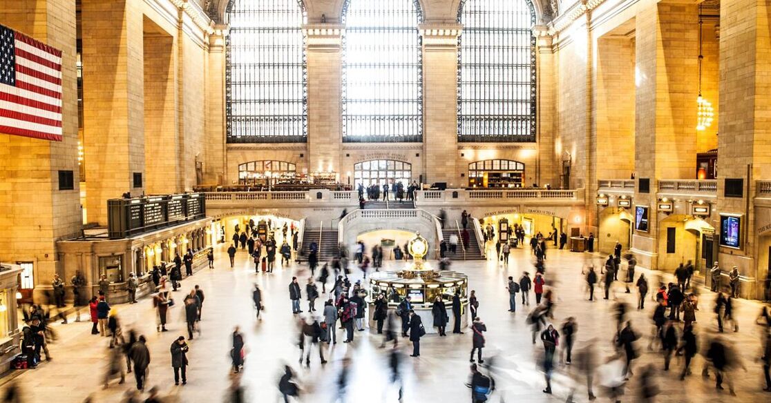 The image shows the bustling main concourse of Grand Central Terminal in New York City, with many people moving about and the American flag hanging from the wall.