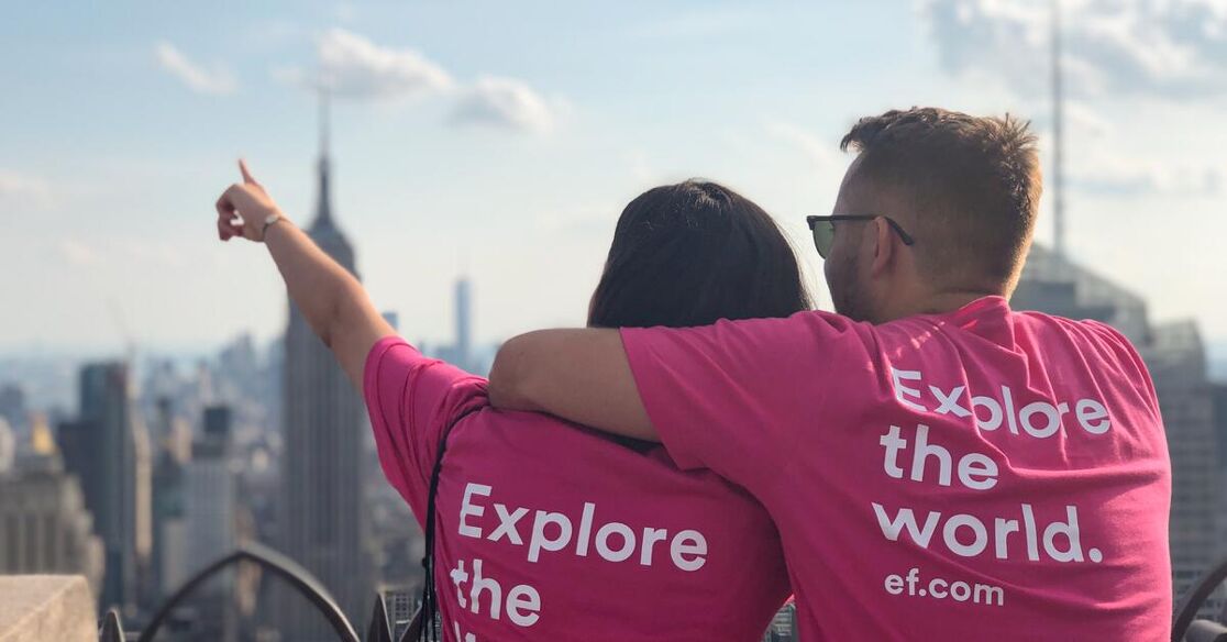 Two people wearing pink t-shirts with "Explore the world. ef.com" printed on them are looking at a cityscape, with one person pointing towards the view.