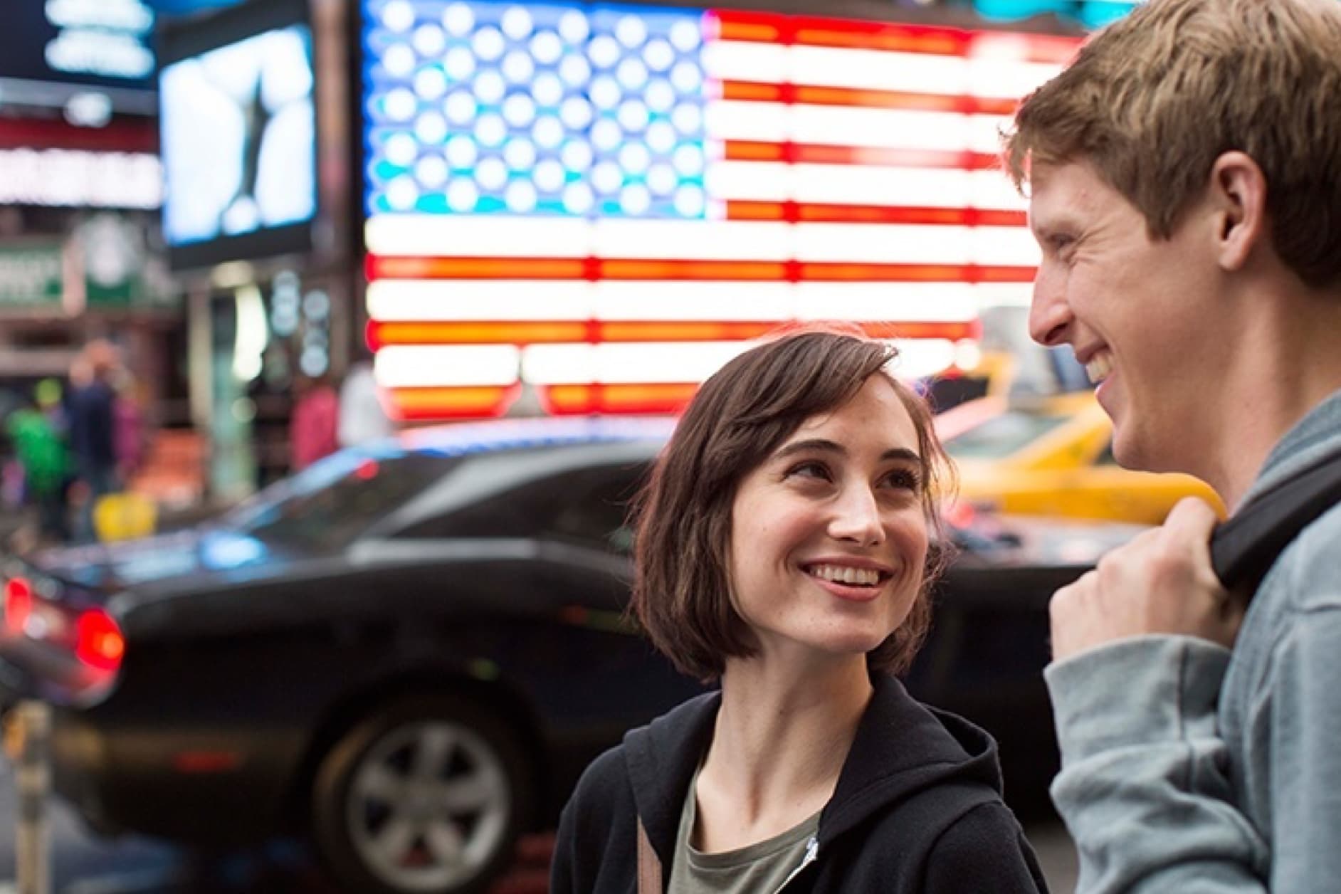 A man and a woman are smiling at each other in a bustling city environment, with a large American flag illuminated on a building in the background.