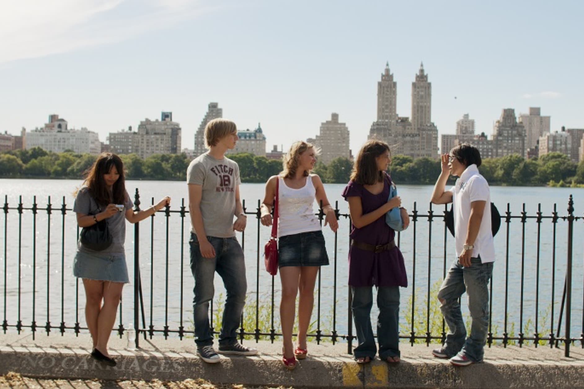 A group of five young adults are standing by a body of water, with a cityscape in the background. They appear to be casually conversing on a sunny day.