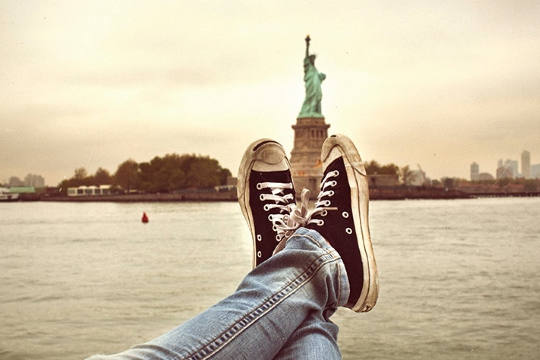 This image captures a unique perspective of the Statue of Liberty, with the viewer's legs and sneakers prominently in the foreground, suggesting a relaxed and personal encounter with the iconic landmark.