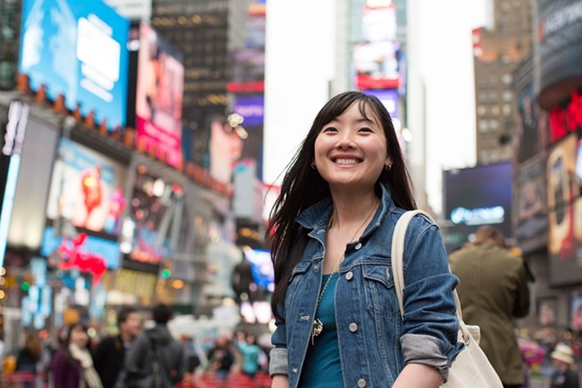 A young Asian woman smiles in the bustling Times Square in New York City, surrounded by bright billboards and crowds of people.