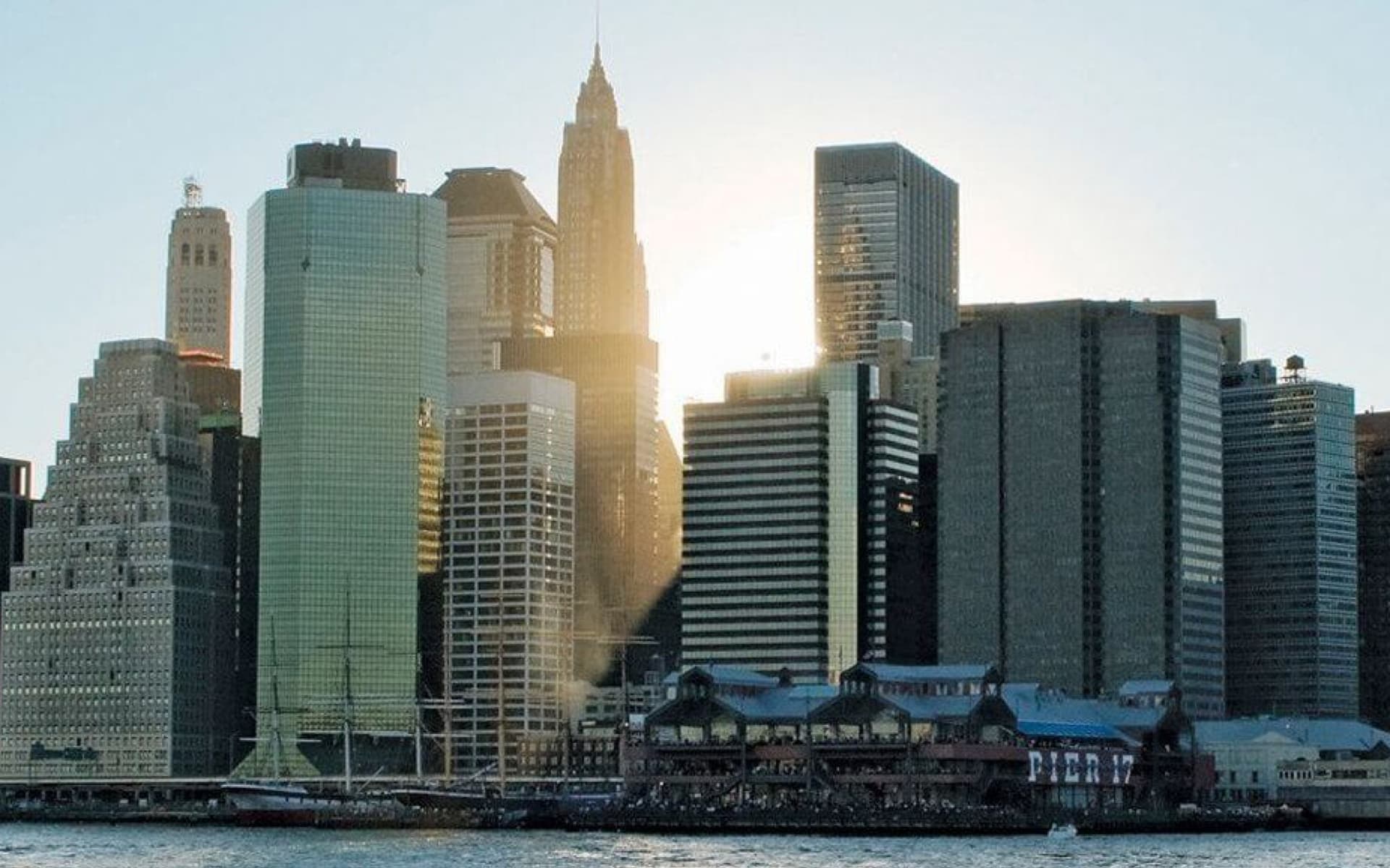 The image shows a cityscape with numerous skyscrapers of varying heights and architectural styles, bathed in the warm glow of the sun peeking from behind the buildings.