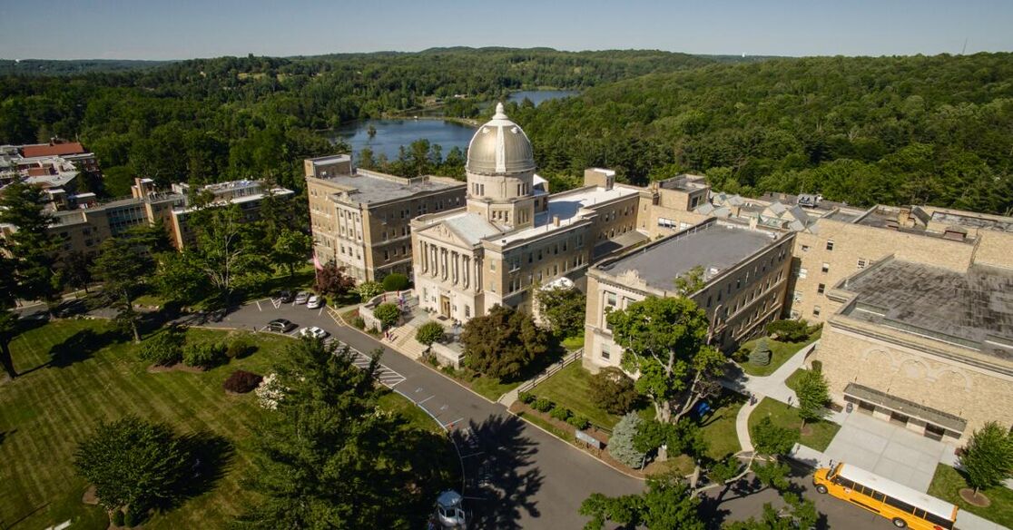 This is an aerial view of a large institutional building with a prominent dome, surrounded by lush green trees and a distant body of water under a clear blue sky.