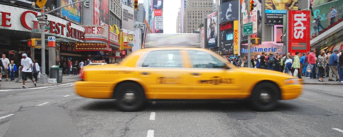 A yellow New York City taxi speeds through the bustling streets of Times Square, surrounded by bright billboards and crowds of people.