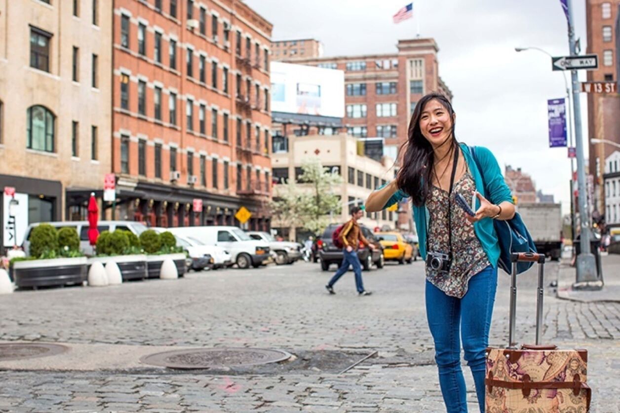 A smiling Asian woman with a suitcase hails a taxi on a cobblestone street in a city, possibly New York, with buildings and traffic in the background.