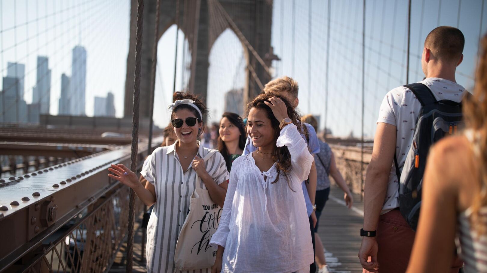 A group of tourists enjoys a sunny day walking across the Brooklyn Bridge in New York City, with the skyline in the background.