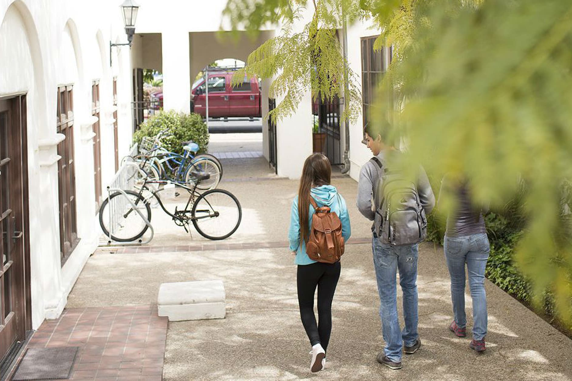 Three students walk down a sun-dappled path between buildings, with bicycles parked nearby. One student in front has a blue hoodie and a brown backpack.