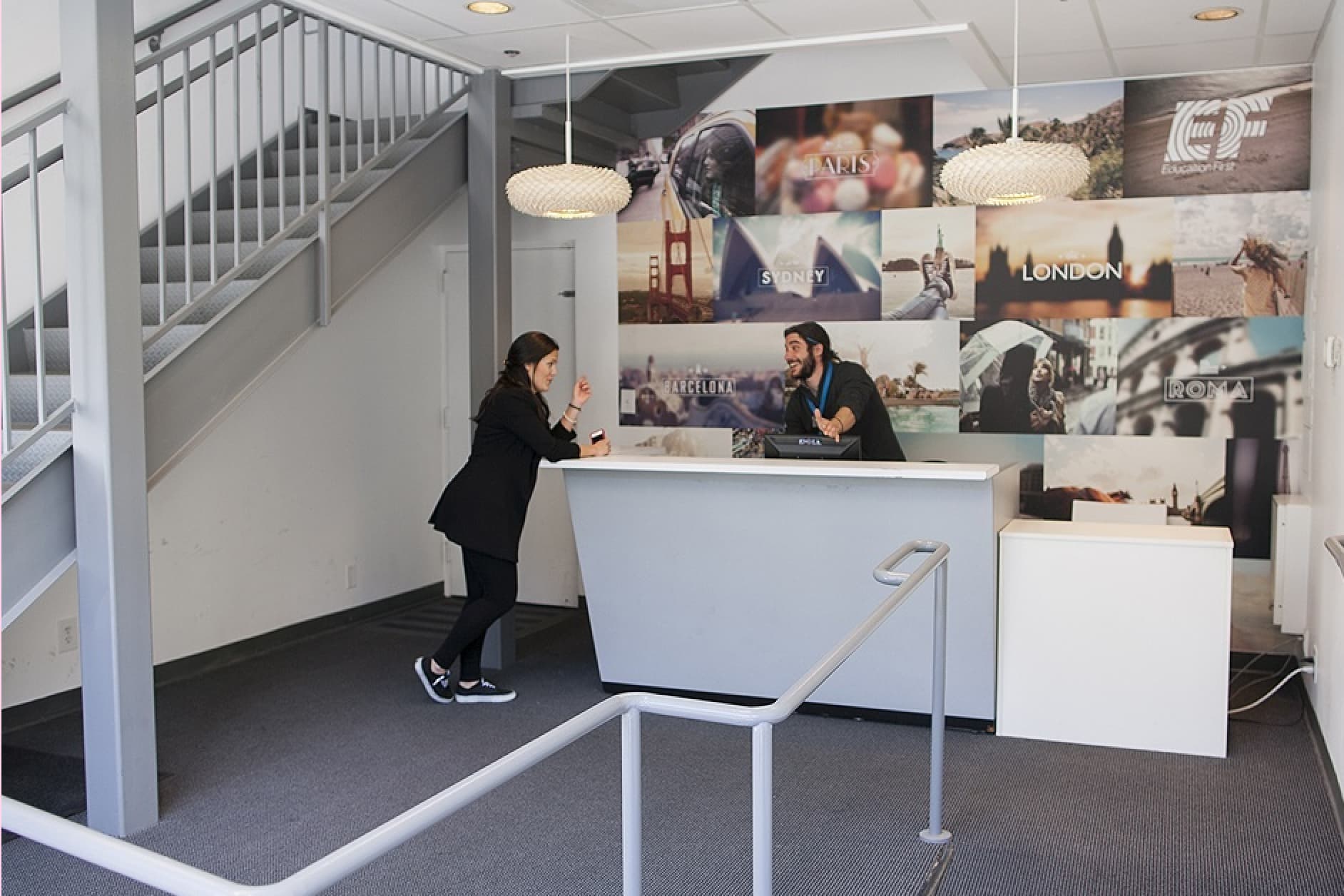 A man and a woman stand at a reception desk in a modern office building, featuring a staircase and a wall collage of landmarks.