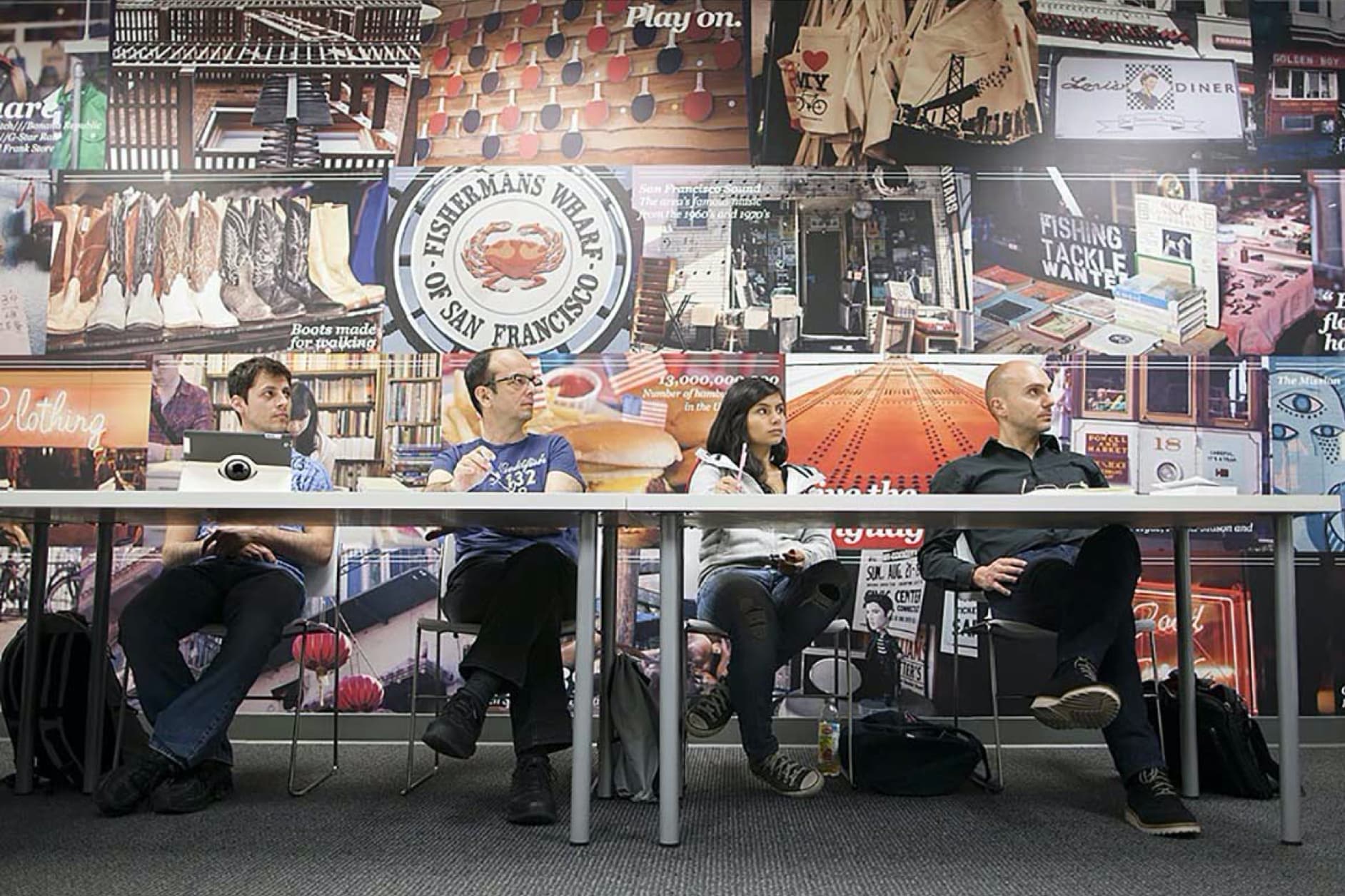 Four people are sitting at a table in front of a wall covered in collages of various images and textures. They appear to be attentive.