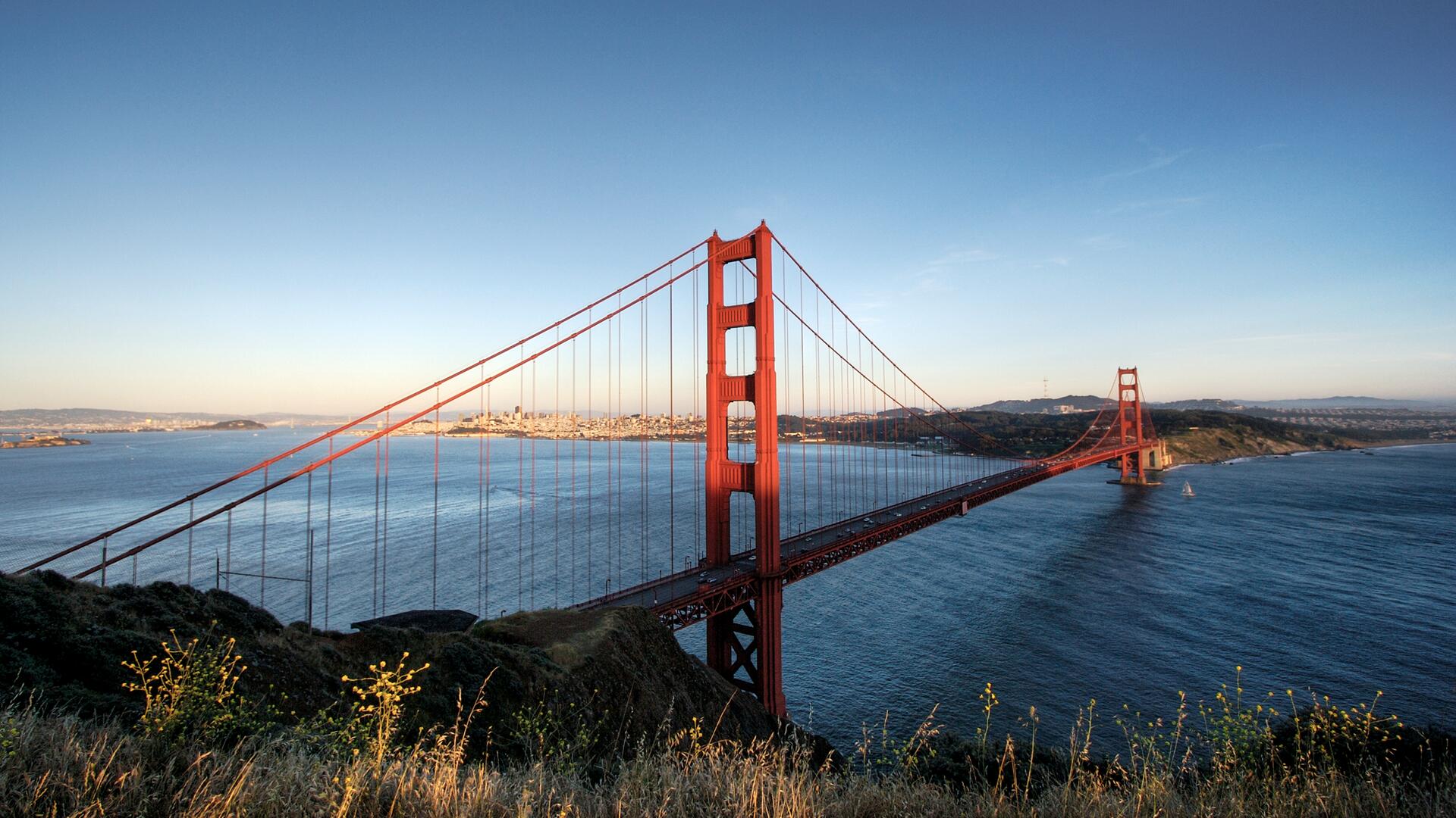 The image shows the Golden Gate Bridge in San Francisco, California, spanning the Golden Gate strait. The iconic red suspension bridge is seen from a high vantage point, with the blue ocean and a cityscape in the background under a clear sky.