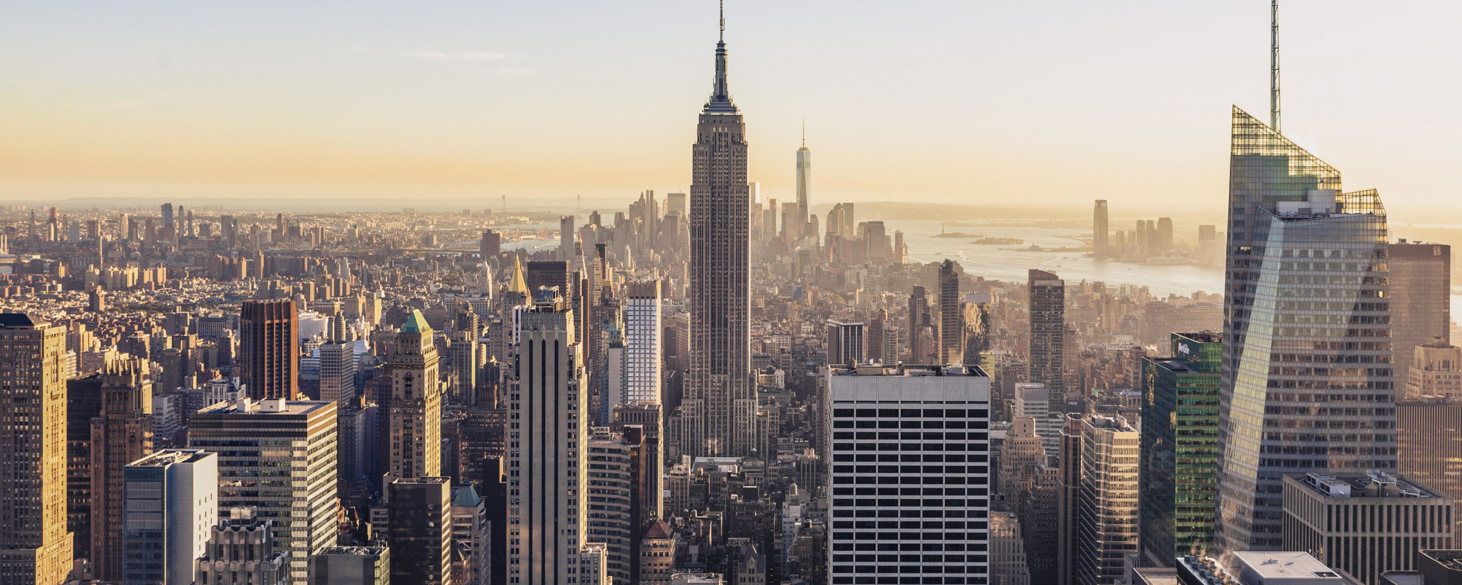 This is a panoramic view of the New York City skyline at sunset, featuring iconic skyscrapers like the Empire State Building and One World Trade Center.