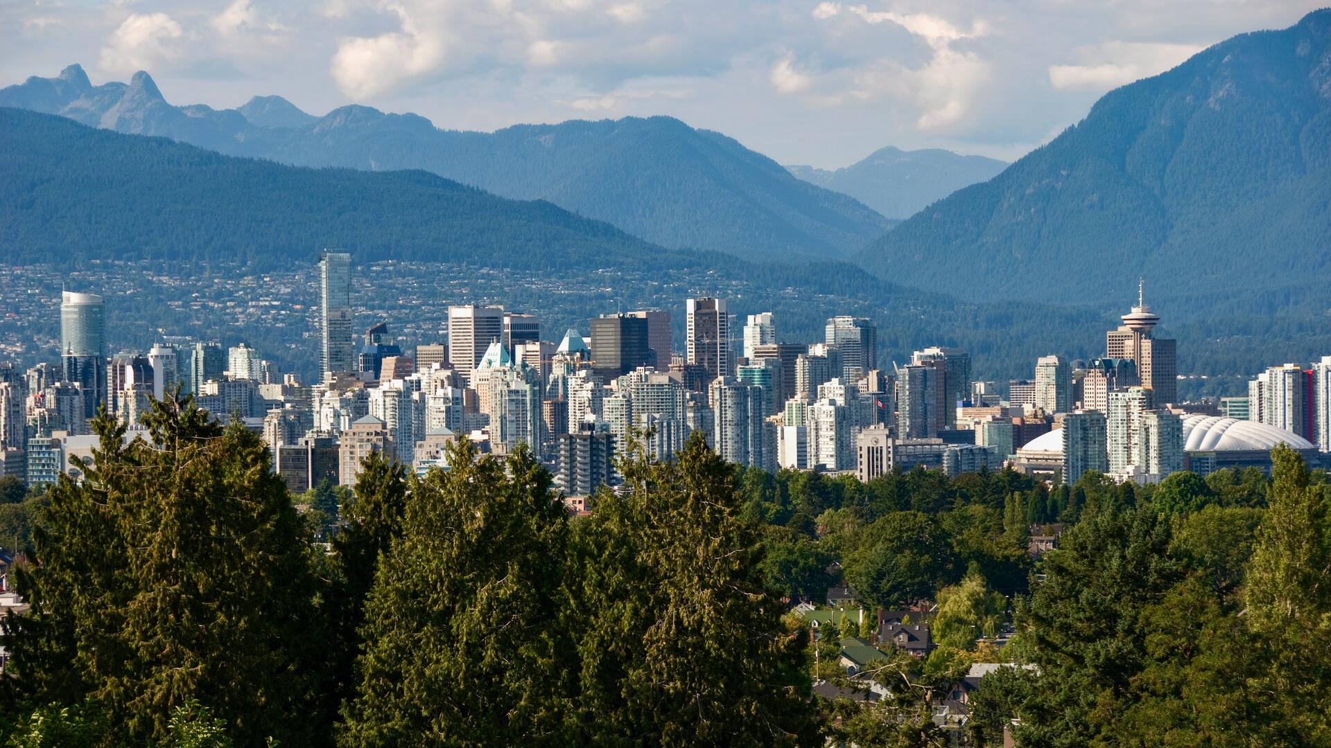 This is a scenic cityscape of Vancouver, Canada, with a dense collection of skyscrapers nestled against a backdrop of majestic, tree-covered mountains under a partly cloudy sky.