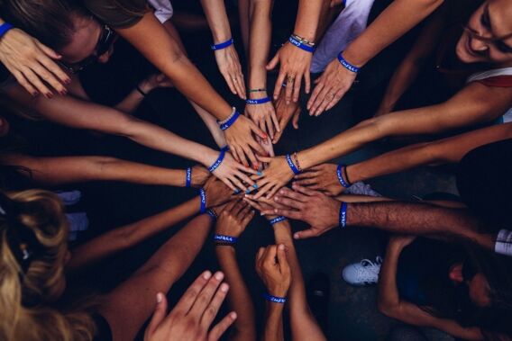 A group of people place their hands in the center, symbolizing teamwork and unity. They are all wearing blue wristbands, suggesting a common cause or event.