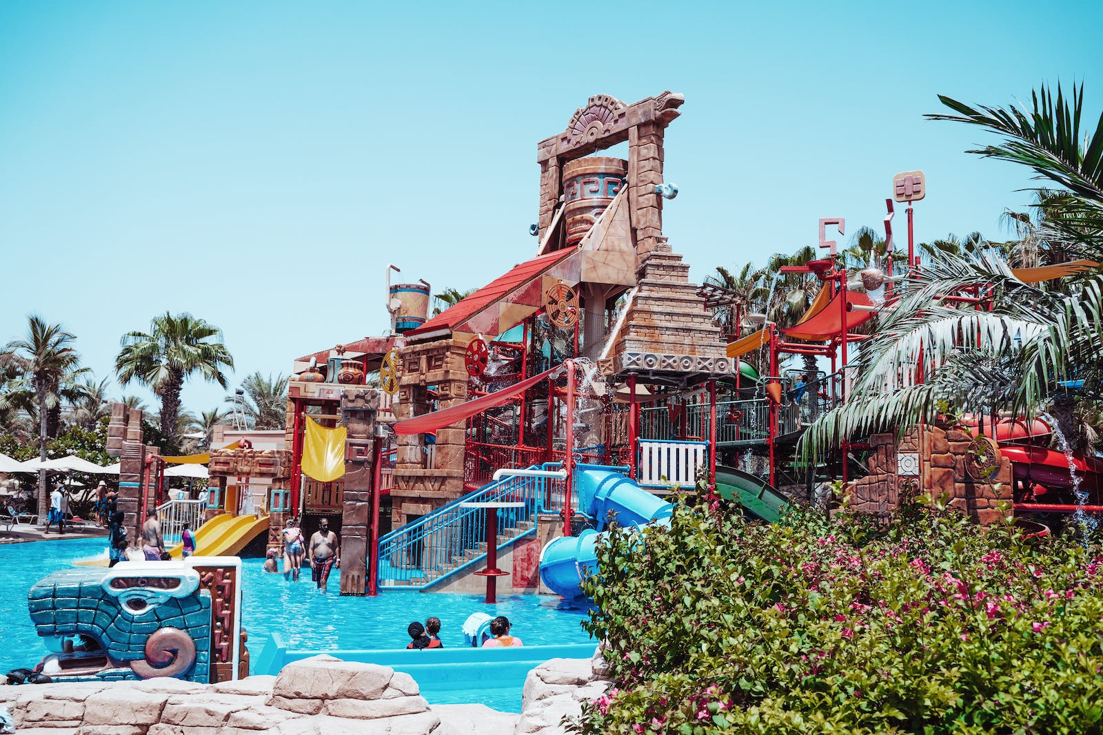 This is a vibrant water park with a large, multi-level play structure featuring slides, water cannons, and a splash pool. Palm trees and lush greenery surround the area, with people enjoying the sunny day.
