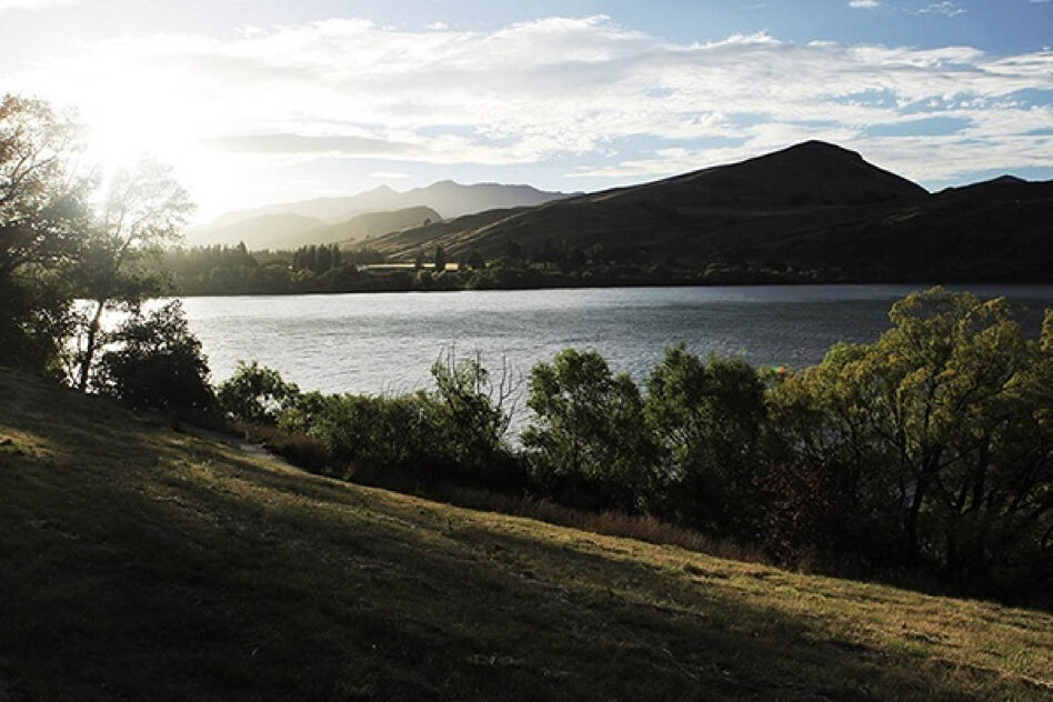 A serene lake is set against a mountainous backdrop as the sun shines through the clouds, casting a warm glow on the landscape. Trees line the shore, and a gentle, grassy hill slopes down in the foreground.
