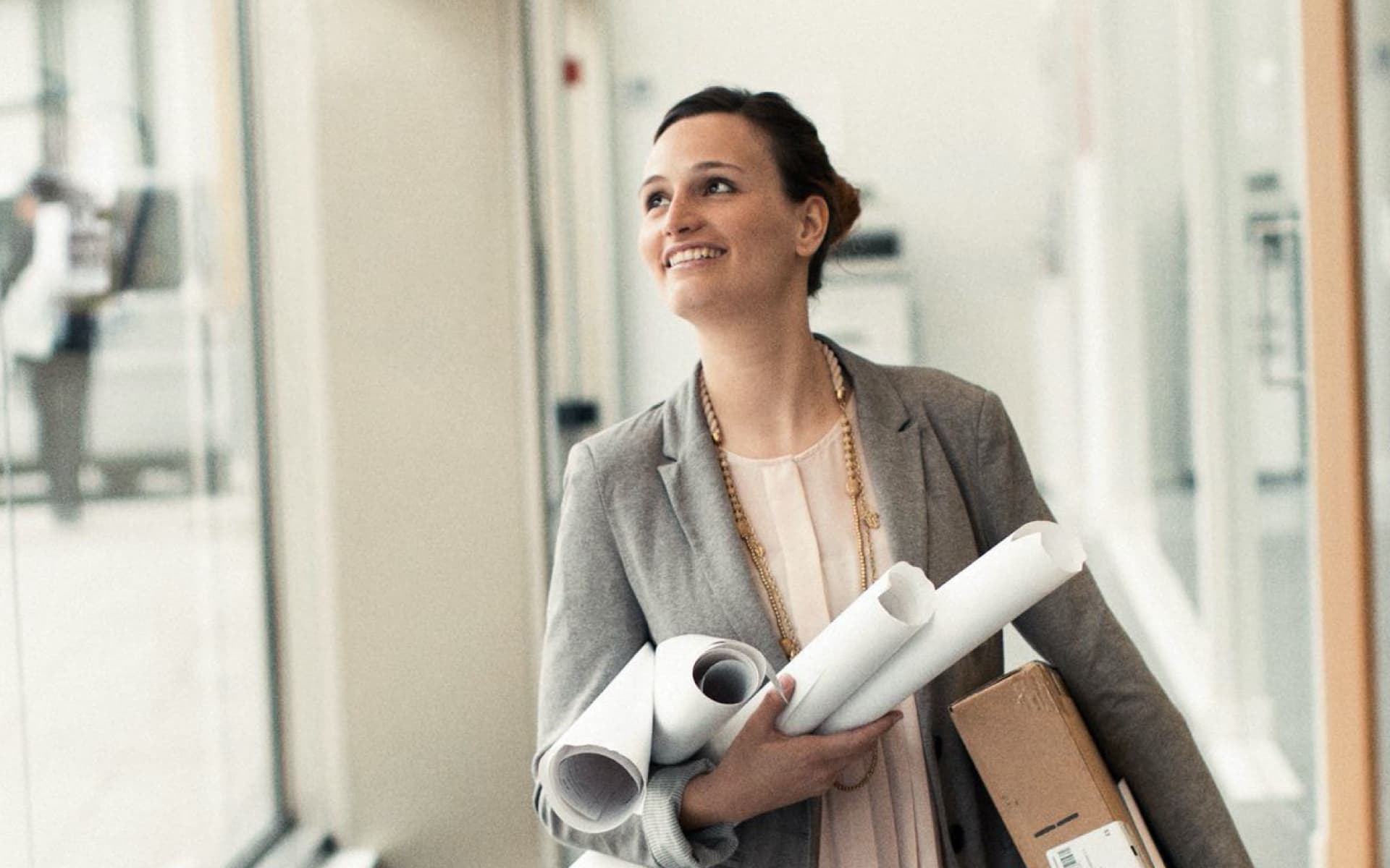 A smiling woman in a grey blazer walks down a hallway carrying blueprints and a file folder, looking towards the light with a confident expression.
