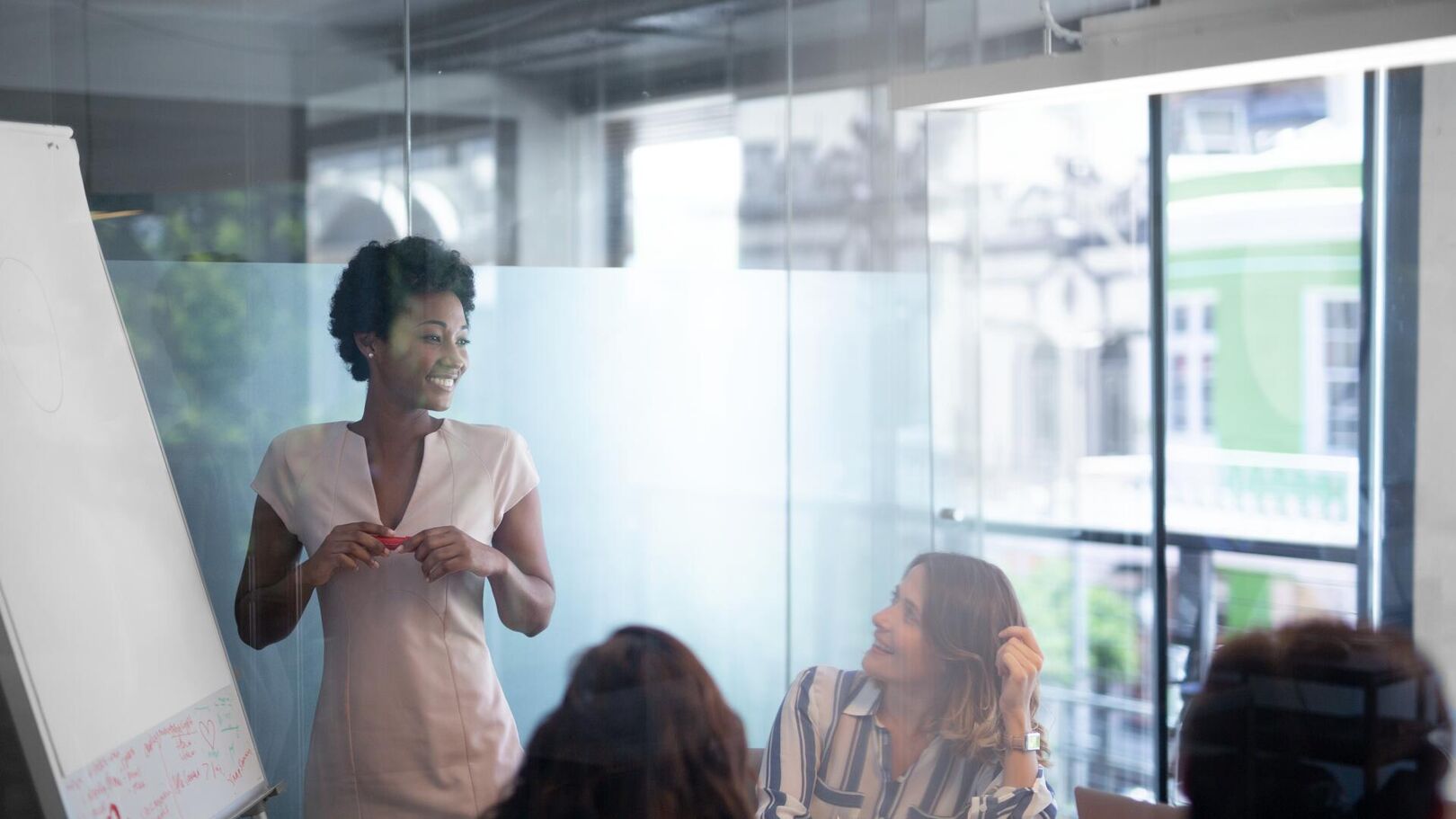 A Black woman is presenting to a diverse group of people in a modern office setting. She is standing next to a whiteboard, holding a marker, and smiling.