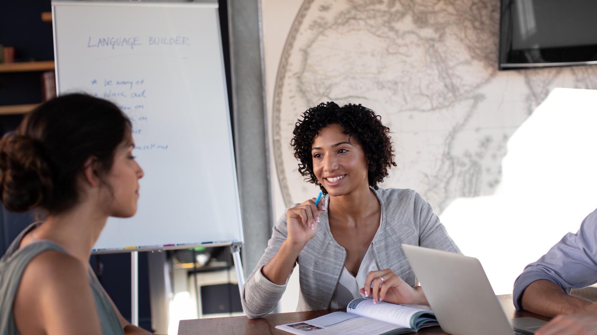 A group of business professionals, including two women and one man, are seated around a table in discussion, with one woman gesturing towards a flip chart.