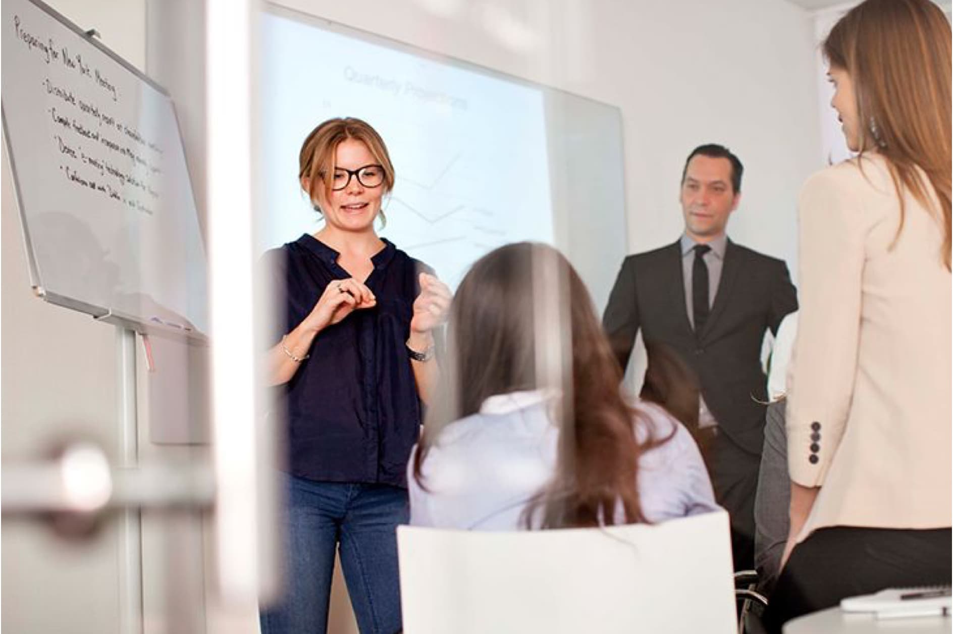 A woman is presenting to a small group of attendees in a modern meeting room, with another woman and a man standing in the background.