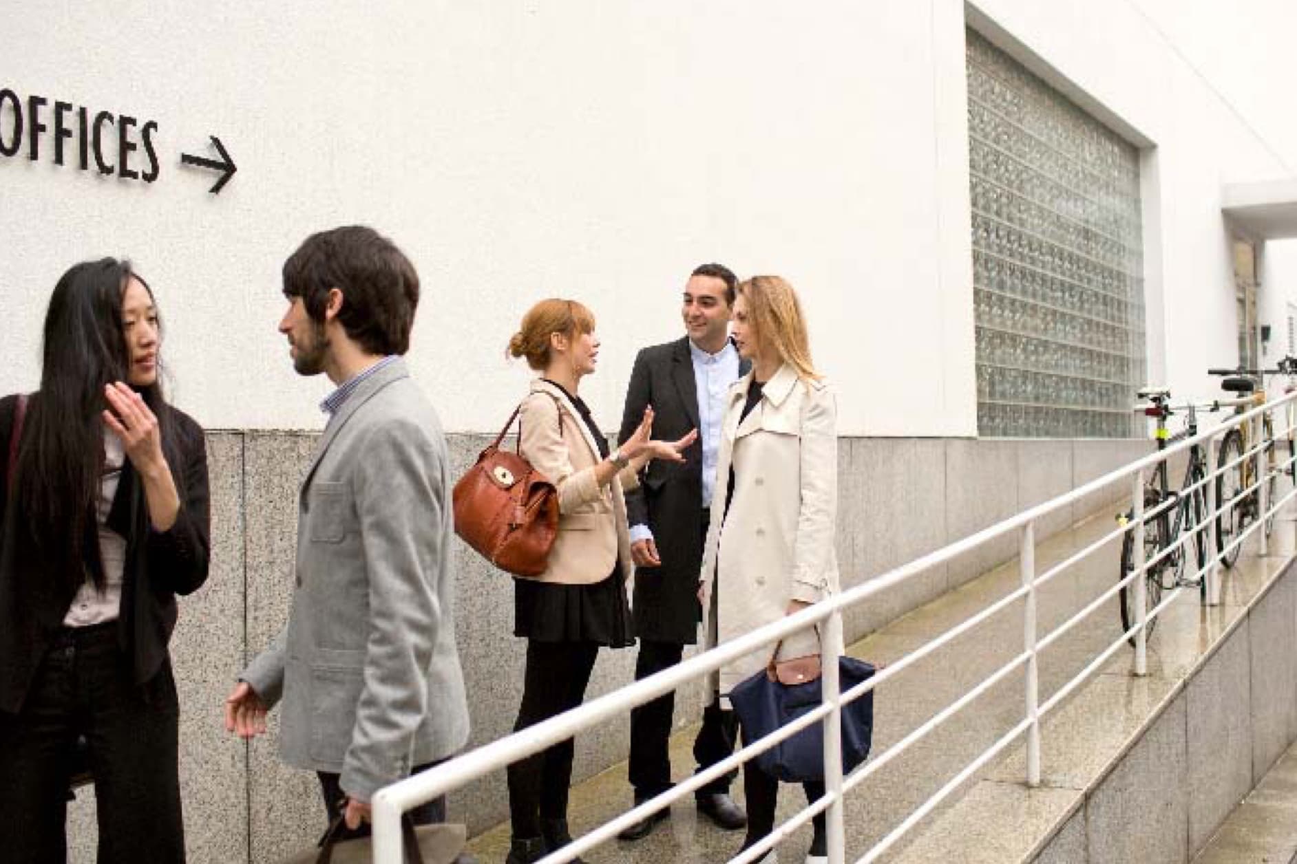 A group of five business people are conversing on a ramp outside an office building.