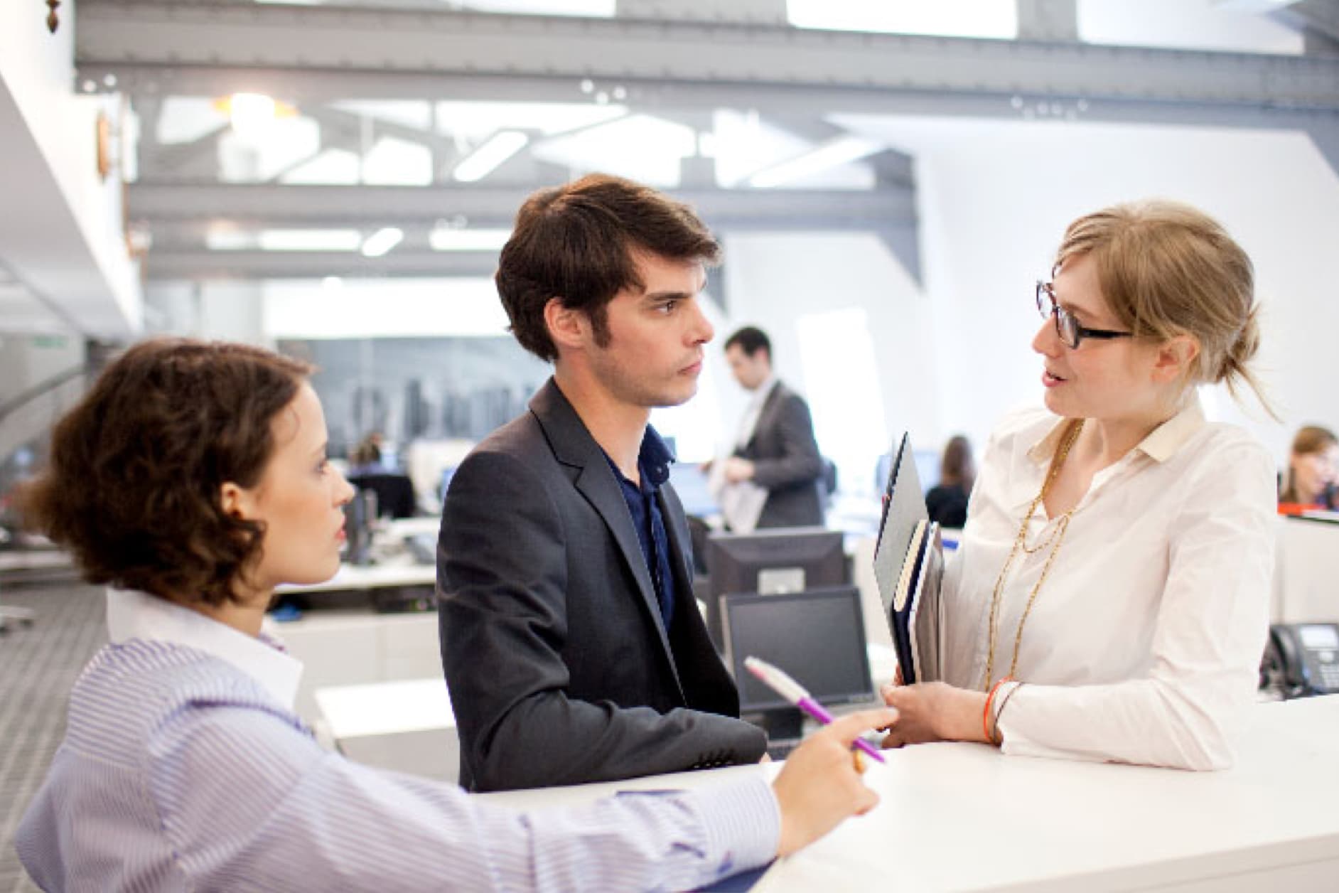 Three people in an office, two women and a man, appear to be conversing at a desk, with others working in the background.