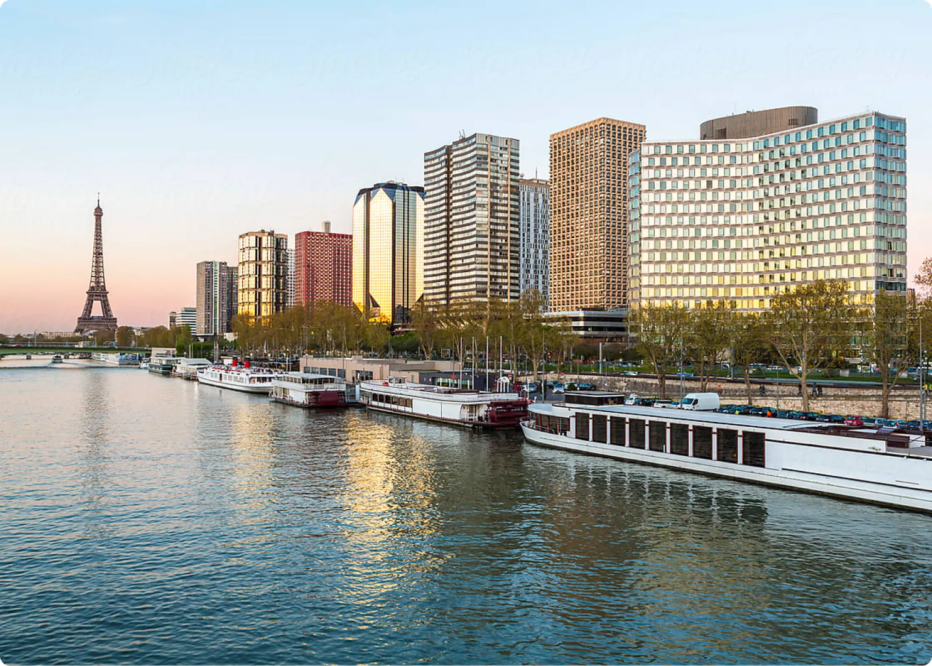 The Seine River flows through Paris, with the Eiffel Tower and modern buildings lining the banks as boats rest on the water.
