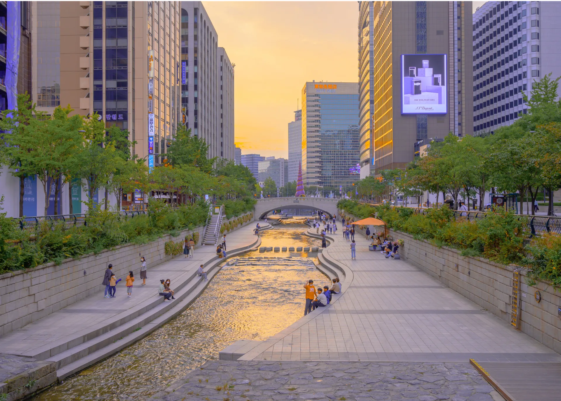 A picturesque view of the Cheonggyecheon Stream in Seoul during sunset, featuring bustling banks, pedestrians, and modern buildings lining the scene.