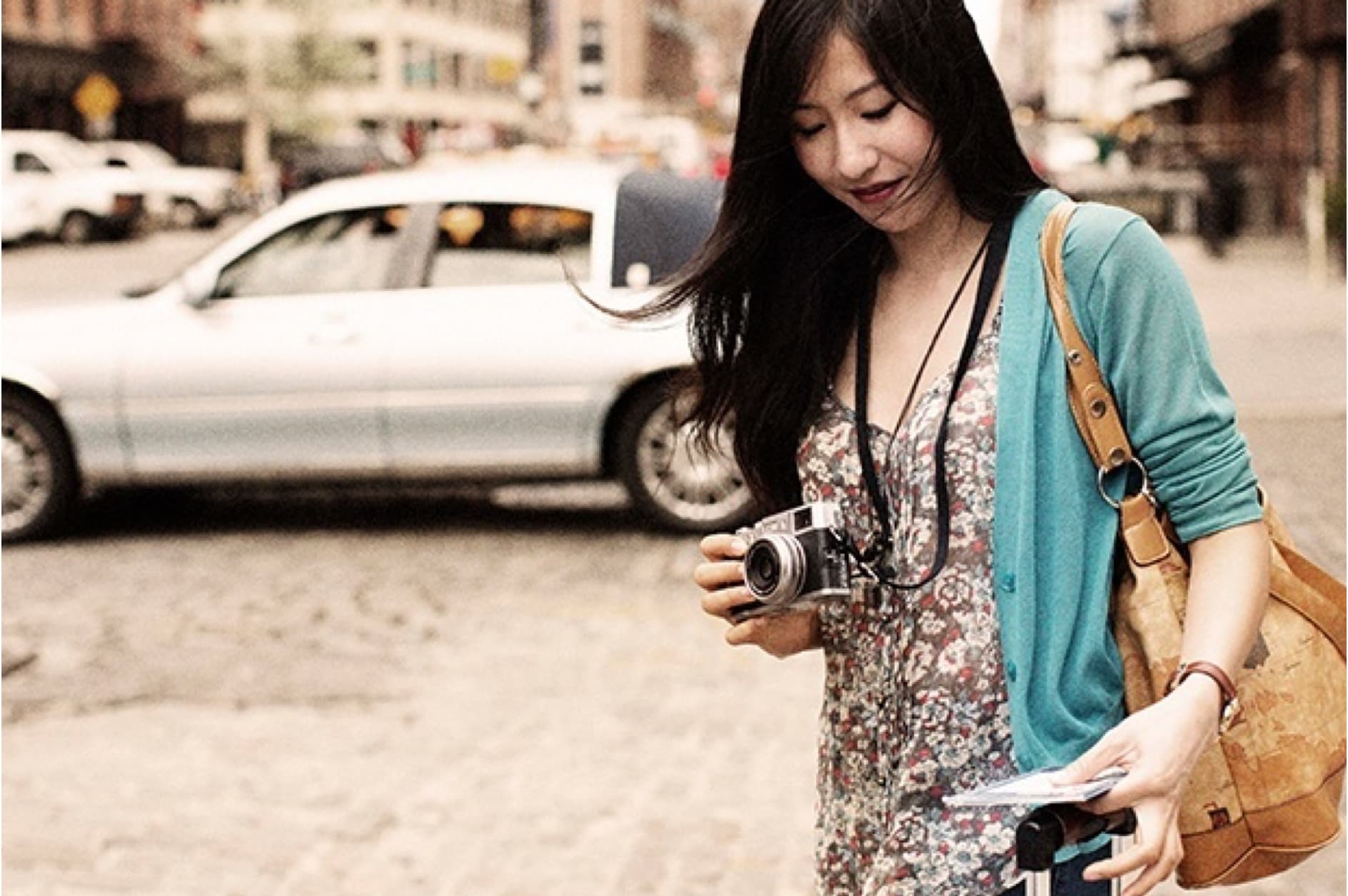 A young Asian woman with long dark hair, wearing a floral dress and a teal cardigan, holds a vintage camera and a map while standing on a cobblestone street with a white limousine in the background.