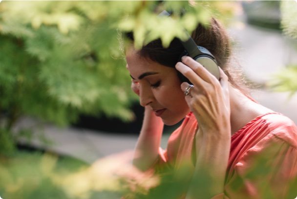 A woman wearing headphones relaxes outdoors, eyes closed, surrounded by lush green foliage, suggesting a moment of peaceful enjoyment and immersion in sound.