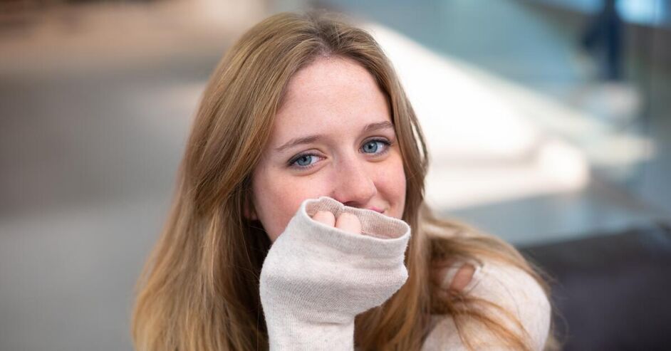 A young blonde girl with freckles and blue eyes smiles, covering her mouth with her sleeve.